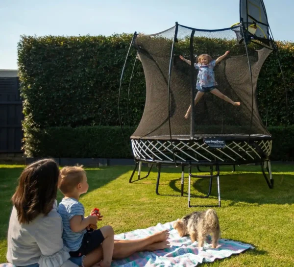 Maminka s malým dítětem sleduje svou dceru skákající na trampolíně Springfree R54 Compact Round